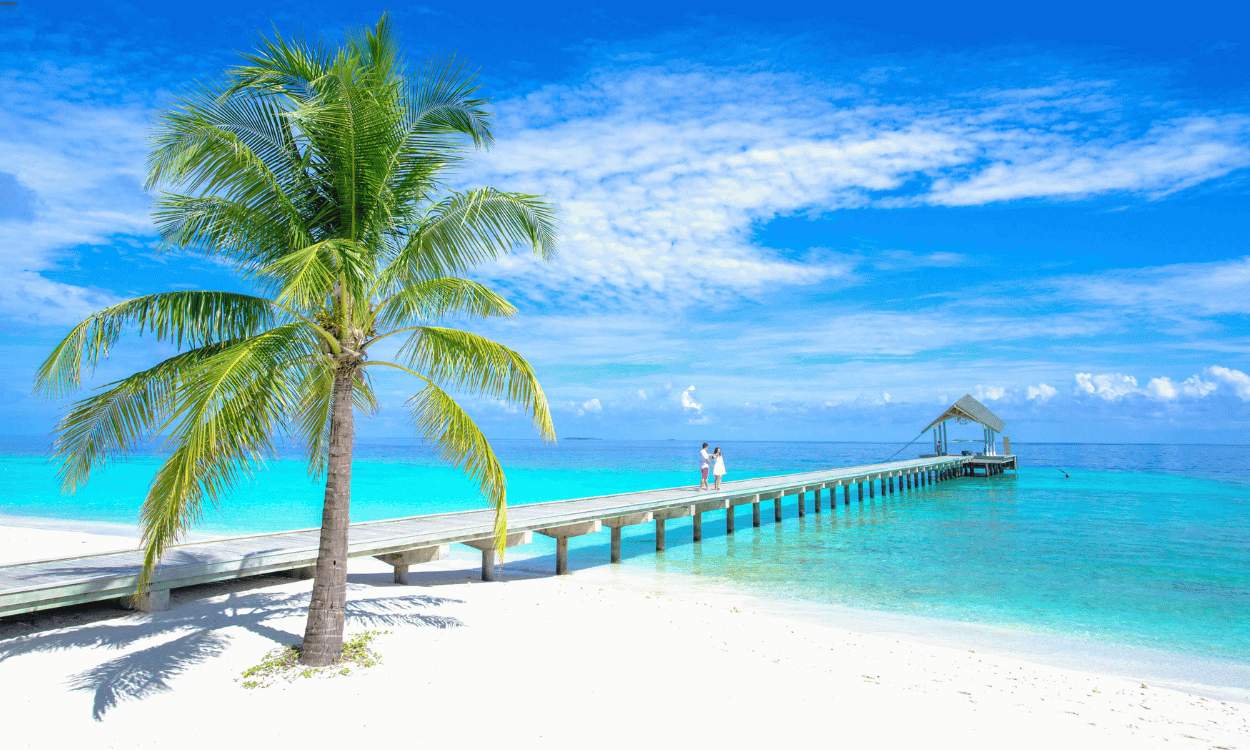 Traumstrand mit weißem Sand und türkisfarbenem Meer. Eine Palme und ein Holzsteg, auf dem ein Paar zu einer überdachten Anlegestelle läuft. Traumstrand mit weißem Sand und türkisfarbenem Meer. Eine Palme und ein Holzsteg, auf dem ein Paar zu einer überdachten Anlegestelle läuft.