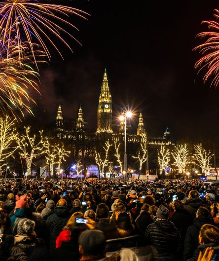 Ein großes Feuerwerk erhellt den Nachthimmel über dem beleuchteten Wiener Rathaus. Davor steht eine dichte Menschenmenge auf einem Platz.